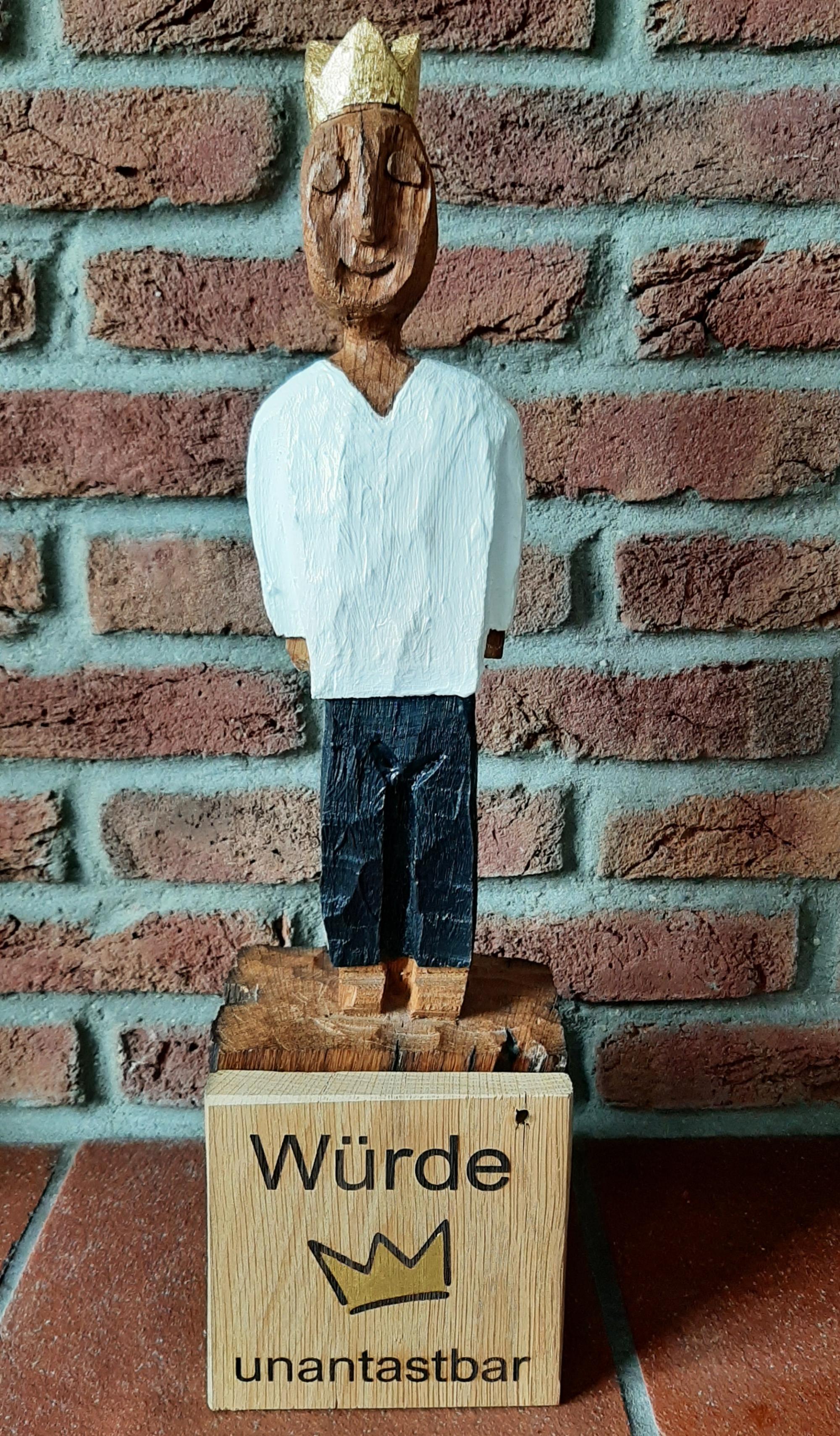 König aus Holz mit einer goldenen Krone. Davor eine Holztafel mit einer Krone und den Wörtern 'Würde unantastbar'.