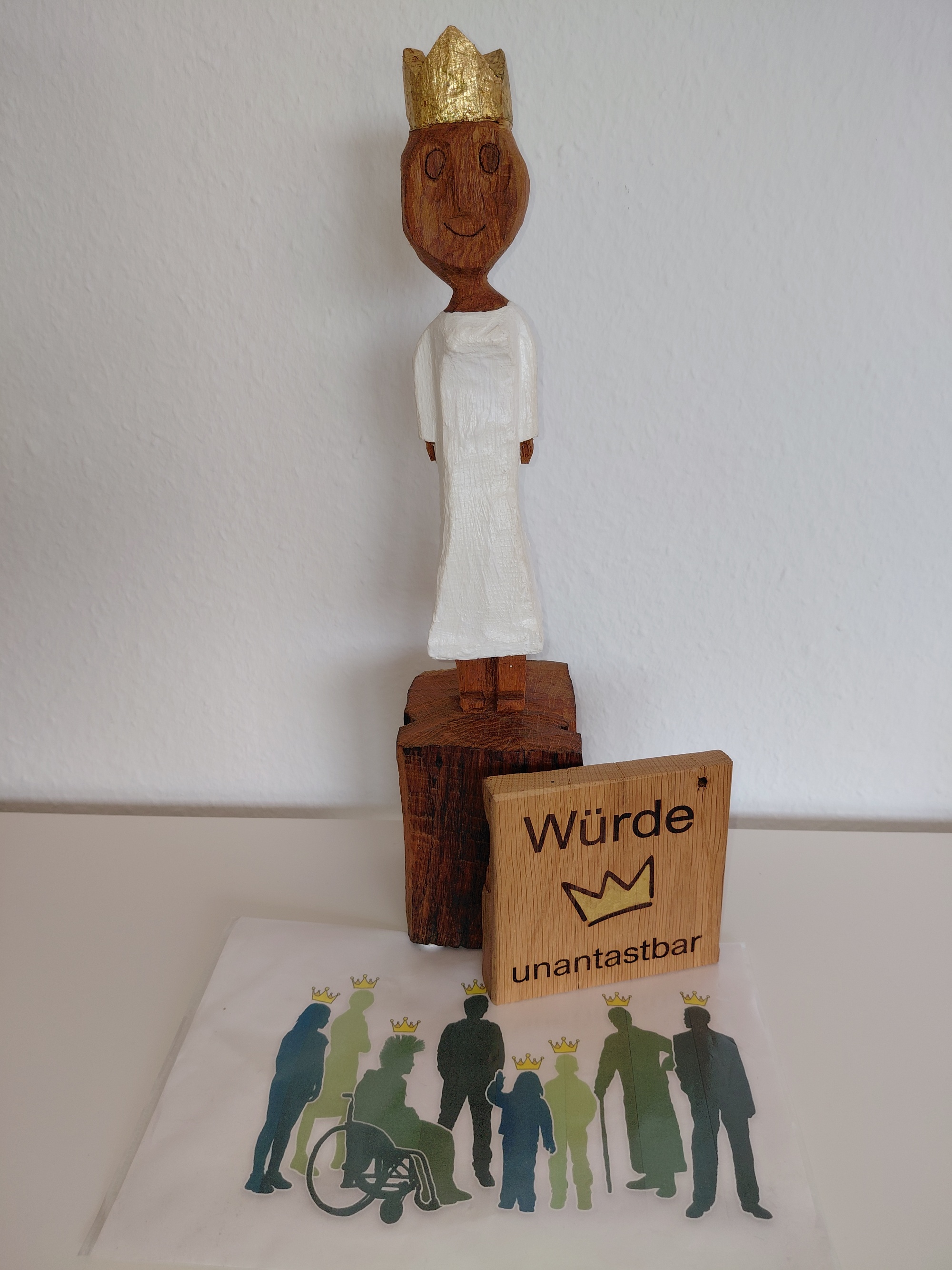 Eine aus Holz geschnitzte Königin. Davor steht eine Holztafel mit einer goldenen Krone und den eingebrannten Wörtern 'Würde unantastbar'. Außerdem noch eine Grafik mit verschiedenen Menschen, die alle die gleiche goldenen Krone tragen