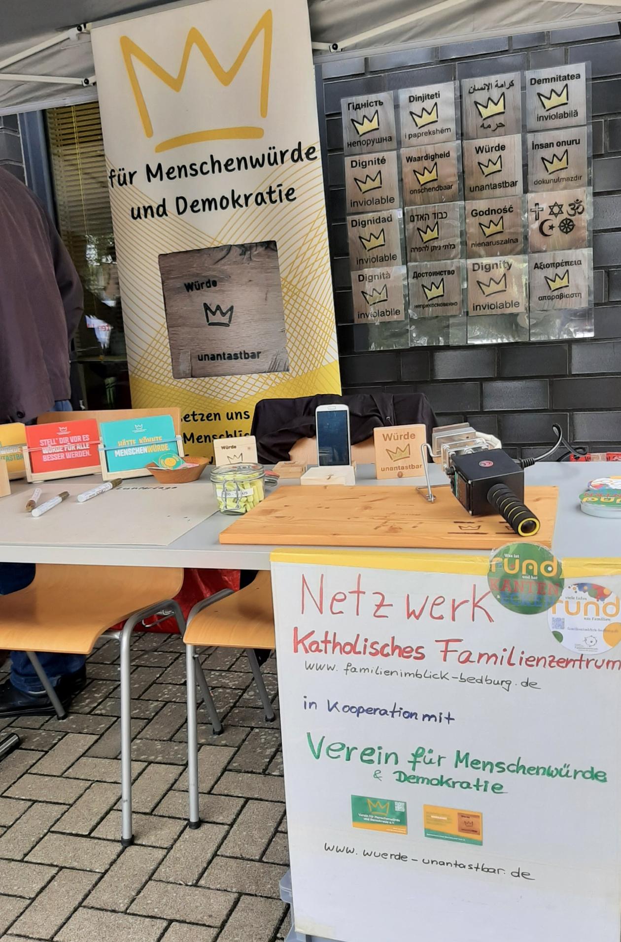 Auf dem Bild ist ein Stand des Netzwerkes Katholisches Familienzentrum in Kooperation mit dem Verein für Menschenwürde und Demokratie zu sehen. Man sieht unter anderem einen Stempel für die Würdetafeln und kleine Holztafeln, wo eine Krone und die Wörter 'Würde unantastbar' drauf zu sehen sind.